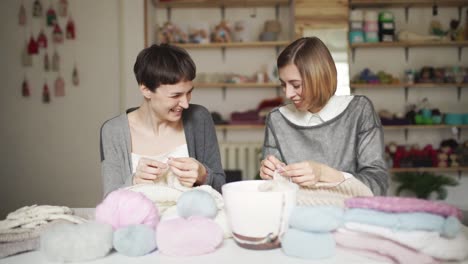 Two-happy-woman-friends-have-fun-knitting-wool-clothes-in-home-studio