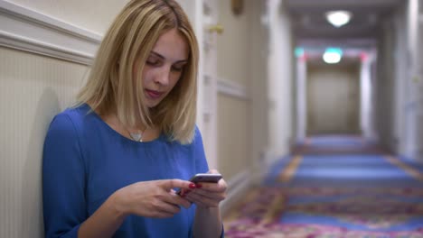 Woman-sitting-on-floor-leaning-on-wall-in-long-corridor-and-using-mobile-phone