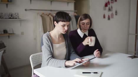 Zwei-Kreative-Frauen,-Die-Ein-Tablet-Für-Ein-Neues-Projekt-Auf-Hängenden-Kleidungsstücken-Im-Hintergrund-Verwenden