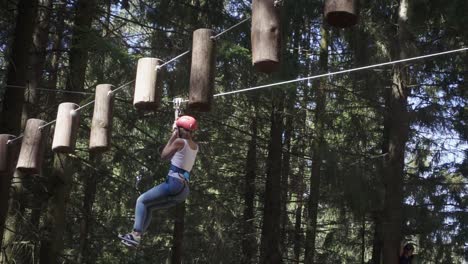 Young-woman-in-safety-equipment-moving-on-rope-between-trees-in-climbing-park