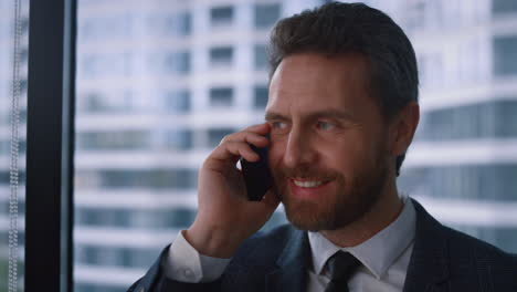 Friendly-businessman-talking-phone-making-corporate-call-in-modern-window-office
