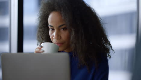 Una-Mujer-Afroamericana-Ejecutiva-Seria-Trabaja-Con-Una-Computadora-Tomando-Café-En-La-Oficina