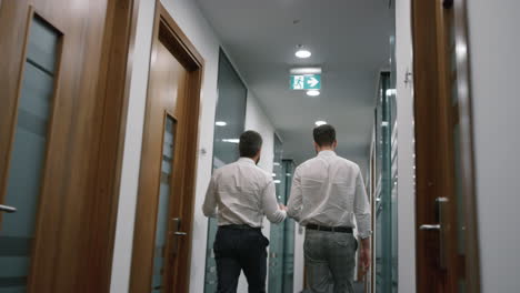 Dos-Hombres-De-Negocios-Caminan-Por-La-Oficina-Discutiendo-Datos-Corporativos-Juntos-En-Una-Compañía-Financiera.