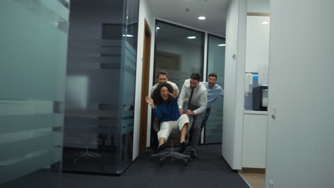 Coworkers-team-enjoy-business-success-ride-office-chair-race-in-diverse-company.