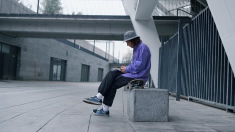 Focused-man-using-smartphone-outdoor.-Young-hipster-sitting-on-skateboard.