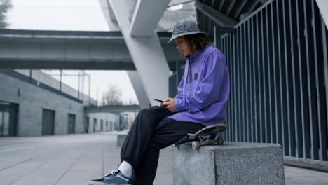 Stylish-man-looking-phone-screen-outdoor.-Skater-typing-message-by-smartphone.