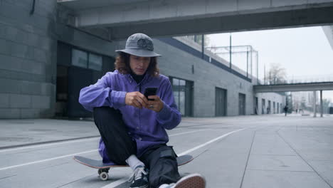 Skater-using-smartphone-outdoor.-Hipster-sitting-on-skateboard-with-phone.