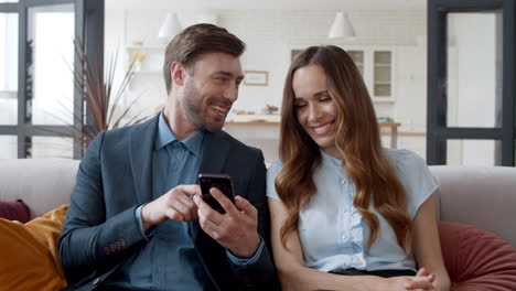 Smiling-couple-looking-mobile-phone-at-home-office.-Laughing-man-and-woman.
