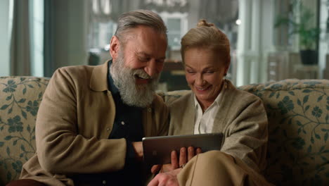 Laughing-mature-couple-looking-tablet-screen-on-sofa-in-classic-living-room.
