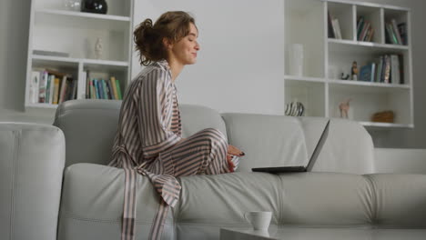 Girl-making-video-call-on-living-room-couch.-Happy-woman-waving-hand-at-laptop