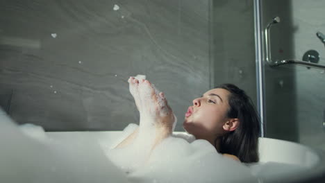 Smiling-girl-blowing-foam-in-tub-in-bathroom.-Beautiful-woman-taking-bubble-bath
