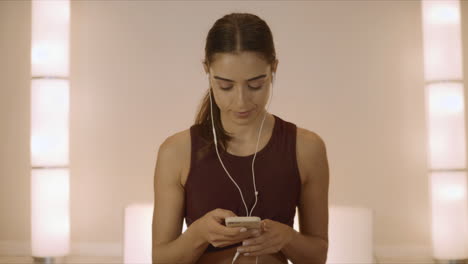 Girl-sitting-in-lotus-pose-on-mat.-Woman-in-earphones-using-smartphone-in-studio