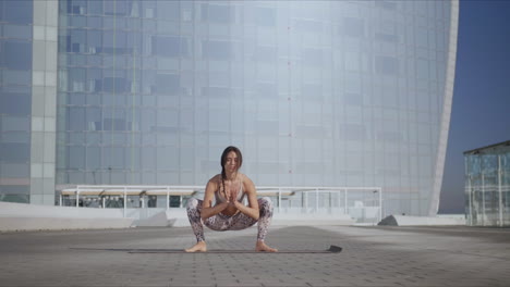 Woman-performing-yoga-in-urban-street.-Lady-doing-garland-pose-on-mat-outdoors