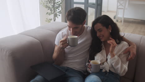 Smiling-couple-drinking-tea-at-home-together.-Sexy-man-and-woman-smiling-at-sofa