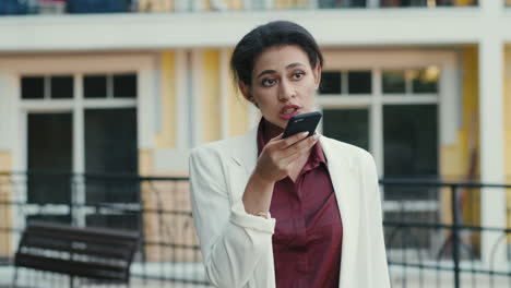 Focused-mixed-race-woman-making-phone-call-outside.-Lady-talking-on-speakerphone