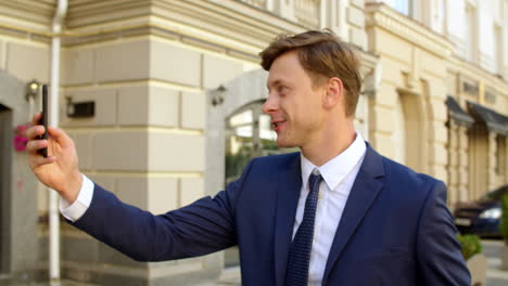 Emotional-business-man-waving-hand-at-video-chat-on-mobile-phone-outdoors
