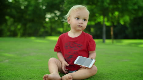 Toddler-sitting-on-grass-in-summer-field.-Closeup-little-boy-holding-smartphone