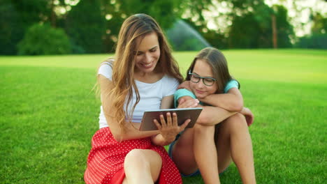Woman-and-girl-sitting-on-grass-with-tablet.-Daughter-and-mother-using-pad