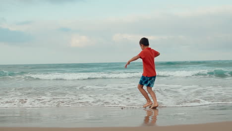Niño-Feliz-Bailando-En-La-Playa-De-Arena-En-Vacaciones.-Chico-Activo-Divirtiéndose-En-El-Surf-En-El-Mar.