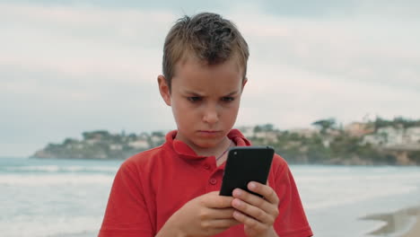 Focused-boy-spending-time-at-beach.-Serious-guy-looking-to-cellphone-at-seaside.