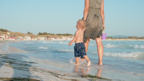Happy-child-looking-around-at-seashore.-Unrecognizable-woman-walking-at-seaside.