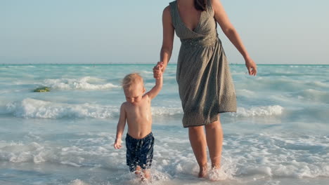 Happy-mother-and-child-going-from-water-at-seashore.-Cute-kid-sitting-in-water.