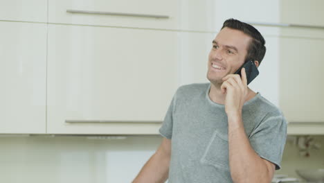Primer-Hombre-Sonriente-Hablando-Por-Teléfono-Móvil-En-La-Cocina-De-Casa.