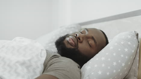 Black-man-sleeping-in-bed-in-morning.-Young-adult-covering-head-with-pillow.