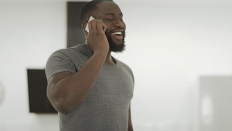 Hombre-Negro-Sonriente-Hablando-Por-Teléfono-En-Casa.-Chico-Joven-De-Pie-Con-Teléfono-Inteligente