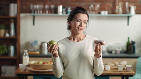 Woman-looking-between-apple-and-eclair.-Brunette-girl-eating-tasty-cake-at-home.