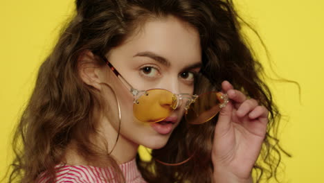 Playful-woman-in-yellow-glasses-looking-to-camera-and-winking-eye-in-studio