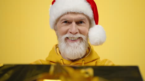 Happy-elderly-man-giving-gift-indoors.-Old-guy-in-santa-hat-posing-in-studio