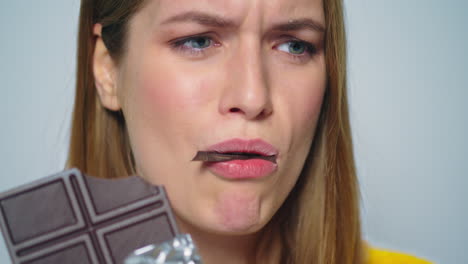 Closeup-joyful-woman-eating-chocolate-bar-at-camera-on-grey-background.