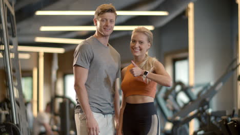 Excited-couple-enjoying-results-in-sport-club.-Fit-girl-showing-like-sign-at-gym