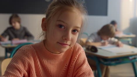 Cute-pupil-sitting-at-desk-at-elementary-school.-Smiling-girl-looking-at-camera