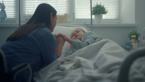 Little-girl-lying-hospital-bed.-Supportive-woman-visiting-holding-child-hand.