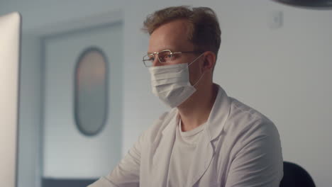 Masked-doctor-watching-monitor-in-clinic-close-up.-Man-physician-writing-report