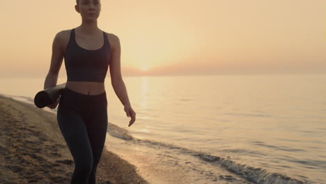 Athletic-woman-walking-beach-holding-sport-mat-at-sunset.-Girl-going-exercising.