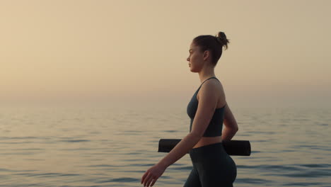 Athlete-girl-walking-seacoast-at-twilight-holding-mat.-Yoga-woman-going-workout.
