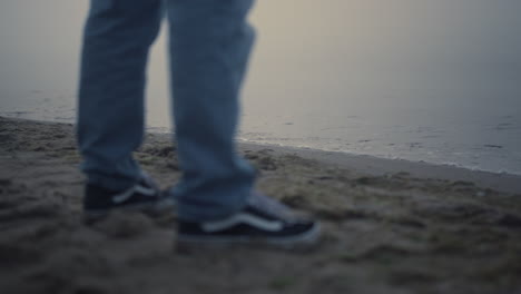 Guy-legs-sneakers-standing-on-sea-beach.-Unknown-man-feet-wearing-casual-shoes