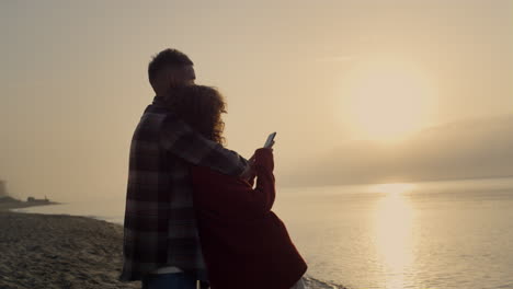 Romantic-couple-looking-sunset-on-beach.-Pretty-woman-taking-photo-of-ocean