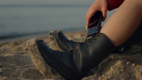 Woman-legs-in-boots-sitting-on-sandy-beach.-Stylish-girl-holding-smartphone