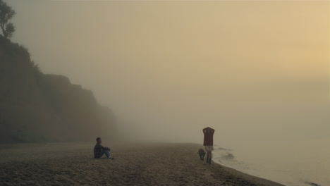 Couple-enjoying-sunrise-on-beach.-Man-sitting-on-shore.-Woman-walking-along-sea