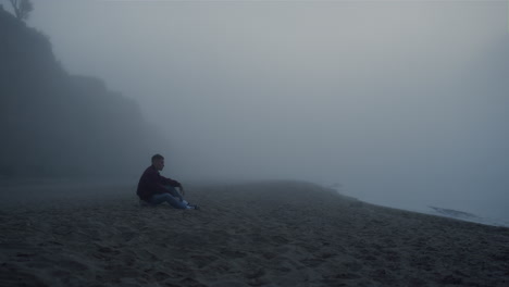 Depressed-guy-sitting-sea-beach-in-foggy-morning.-Worried-man-touching-sand