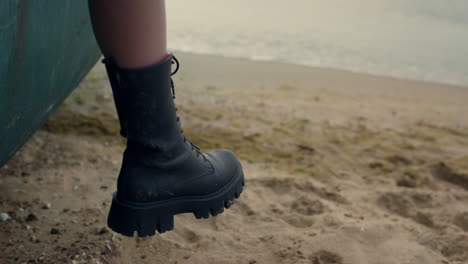 Woman-leg-wearing-black-boot-hanging-from-old-boat-standing-sand-beach-close-up.