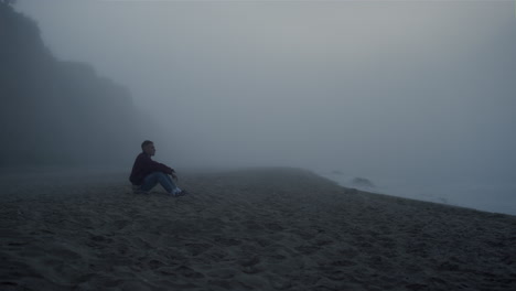 Sad-man-looking-sea-landscape-in-fog.-Serious-guy-sitting-on-sandy-beach