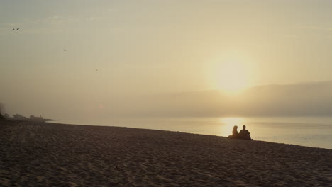 Young-couple-resting-on-beach-at-sunset.-Wide-shot-lovers-dating-at-ocean-coast