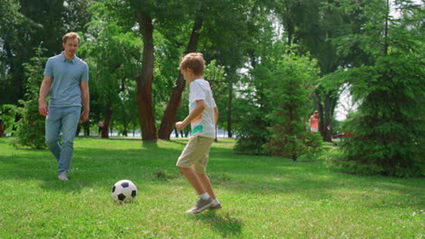 Sporty-man-passing-ball-to-active-son-on-green-park.-Father-play-soccer-with-boy