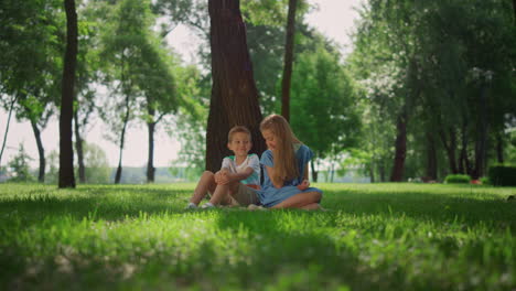 Smiling-siblings-play-on-grass-under-tree.-Happy-children-sitting-on-lawn.