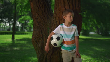 Little-boy-hold-soccerball-lean-on-tree.-Young-athlete-posing-with-ball-closeup.